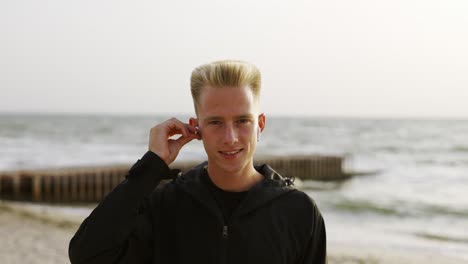 Portrait-of-a-young-man-who-puts-and-earphone-into-his-ear-and-listens-to-music-against-the-backdrop-of-the-waves-of-the-sea-at