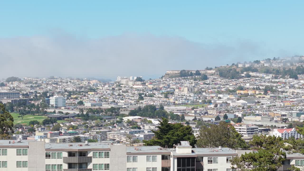 A quiet day unfolds above Broadmoor, where homes and roadways stretch toward Daly City’s coastal hills. Static aerial shot.