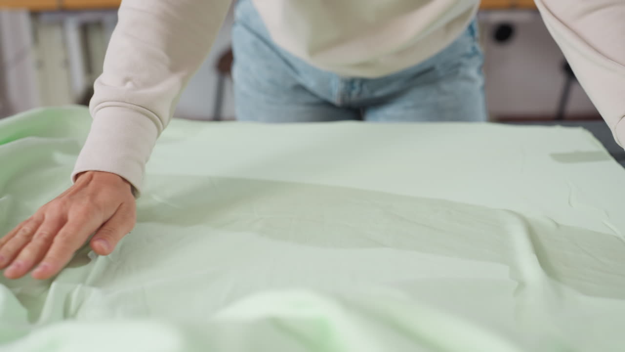 Dress maker stretches light green fabric across table, gently running hands over material to smooth wrinkles, preparing textile in bright sewing workspace with focus on quality and craftsmanship