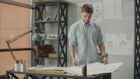 bearded man hipster standing faces to panoramic window and looks at blueprints. young architect in stylish loft office works with documents. creative architect thinking on the big drawings.