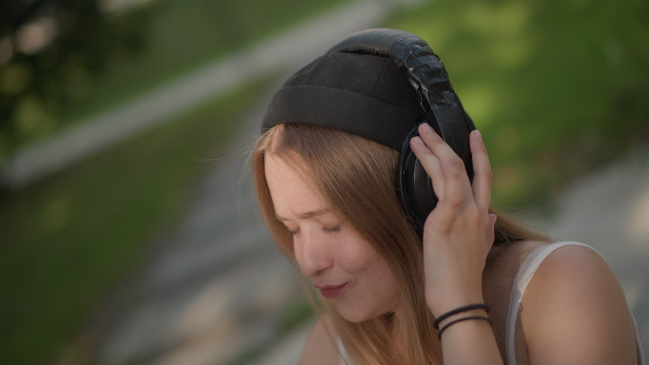 Giovane donna che balla ascoltando musica sul marciapiede, cuffie, occhi chiusi e sorriso, gesti ritmici delle mani e leggero ondeggiamento, luce del sole sui capelli e sfondo del parco, umore energico e giocoso, espressivo urbano