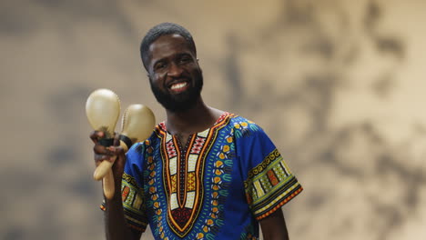 young cheerful african american man in traditional clothes playing maracas and smiling at camera