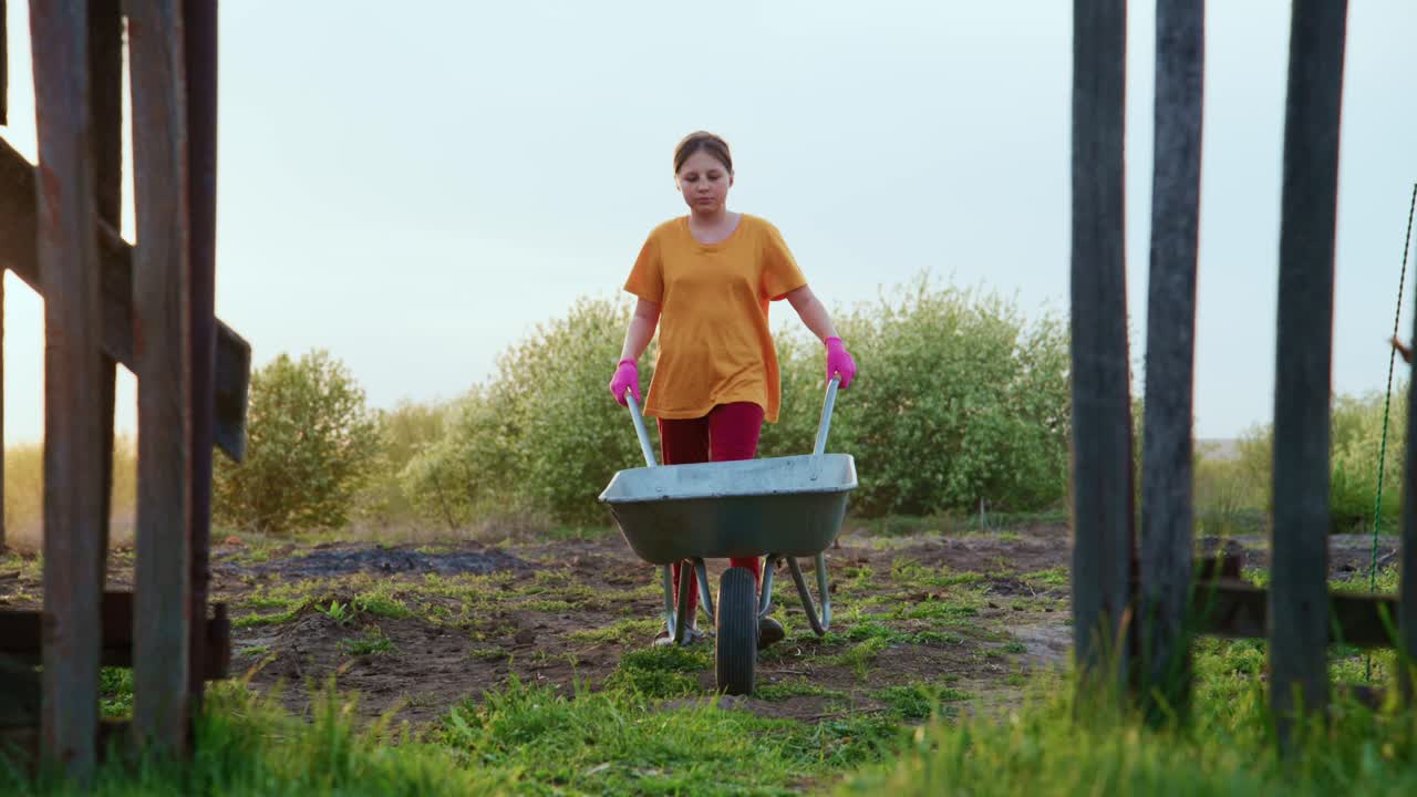Woman pushes wheelbarrow through garden path framed by wooden fence. She wears yellow shirt and pink gloves. Ground is muddy, grassy with trees in background