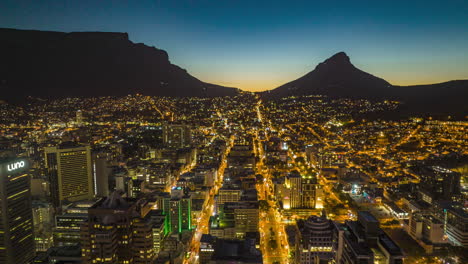 Forwards-fly-above-illuminated-streets-of-city-at-dusk.-Aerial-hyperlapse-footage-of-town-development-with-table-mountain-in-distance.-South-Africa