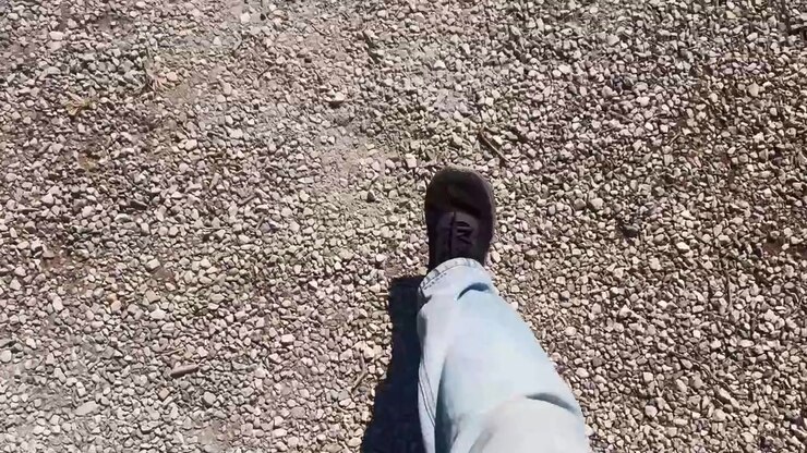 Siyah ayakkabı, açık renk kot pantolon, gevşek taşlar, kuru zemin ve dış mekan seyahat hareketi ile çakıllı yolda yürüyen bir kişinin bakış açısı çekimi