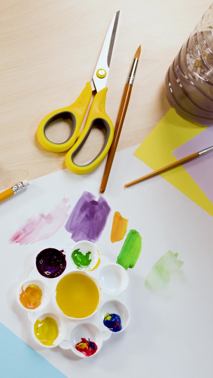 A top-down shot pans across a desk with scissors, brushes, and vibrant watercolor paints