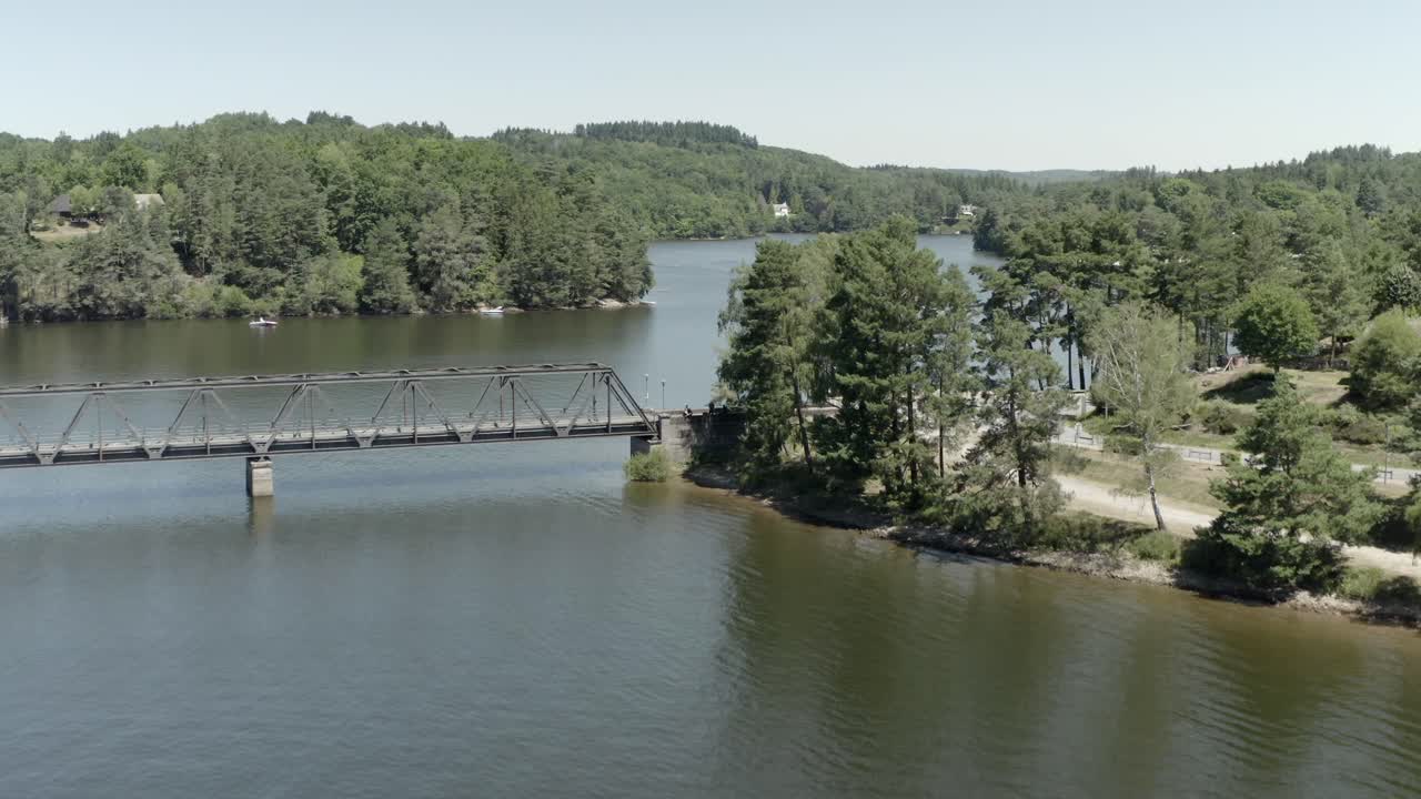 Aerial view rising over calm lake, revealing Pont de Lantourne bridge and forested shoreline, Marcillac La Croisille, France