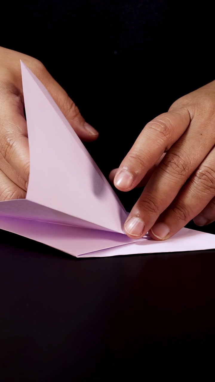 Hænder folder forsigtigt pastelpapir på sort overflade, trin-for-trin origami-forberedelse, nærbillede.