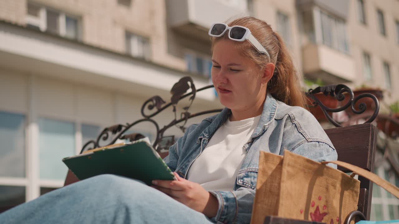 Young woman reading green folder outdoors, relaxed student sits on ornate metal bench near apartment building, wearing denim jacket and sunglasses, headband in hair, paper bag beside her, flipping