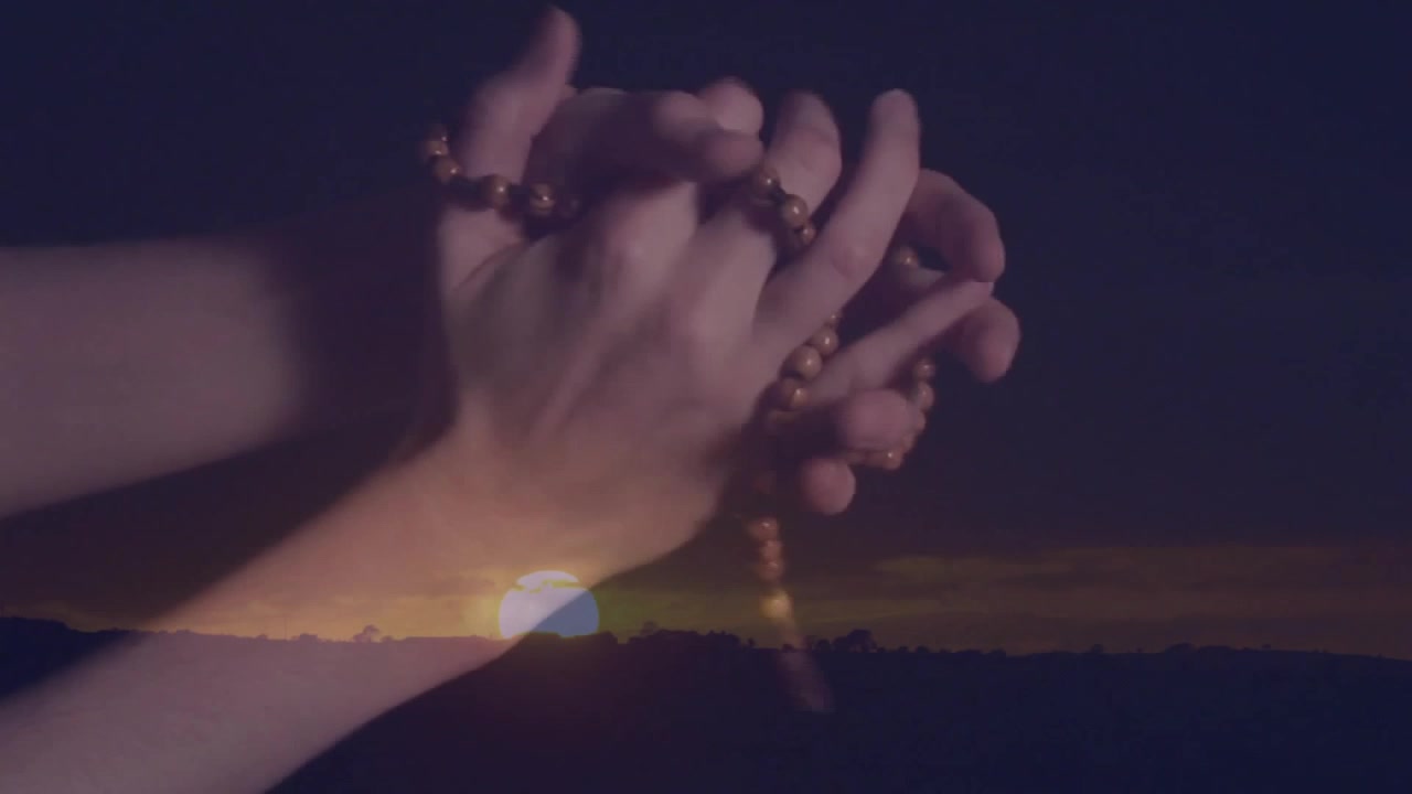Holding rosary beads, hands silhouetted against sunset in peaceful landscape