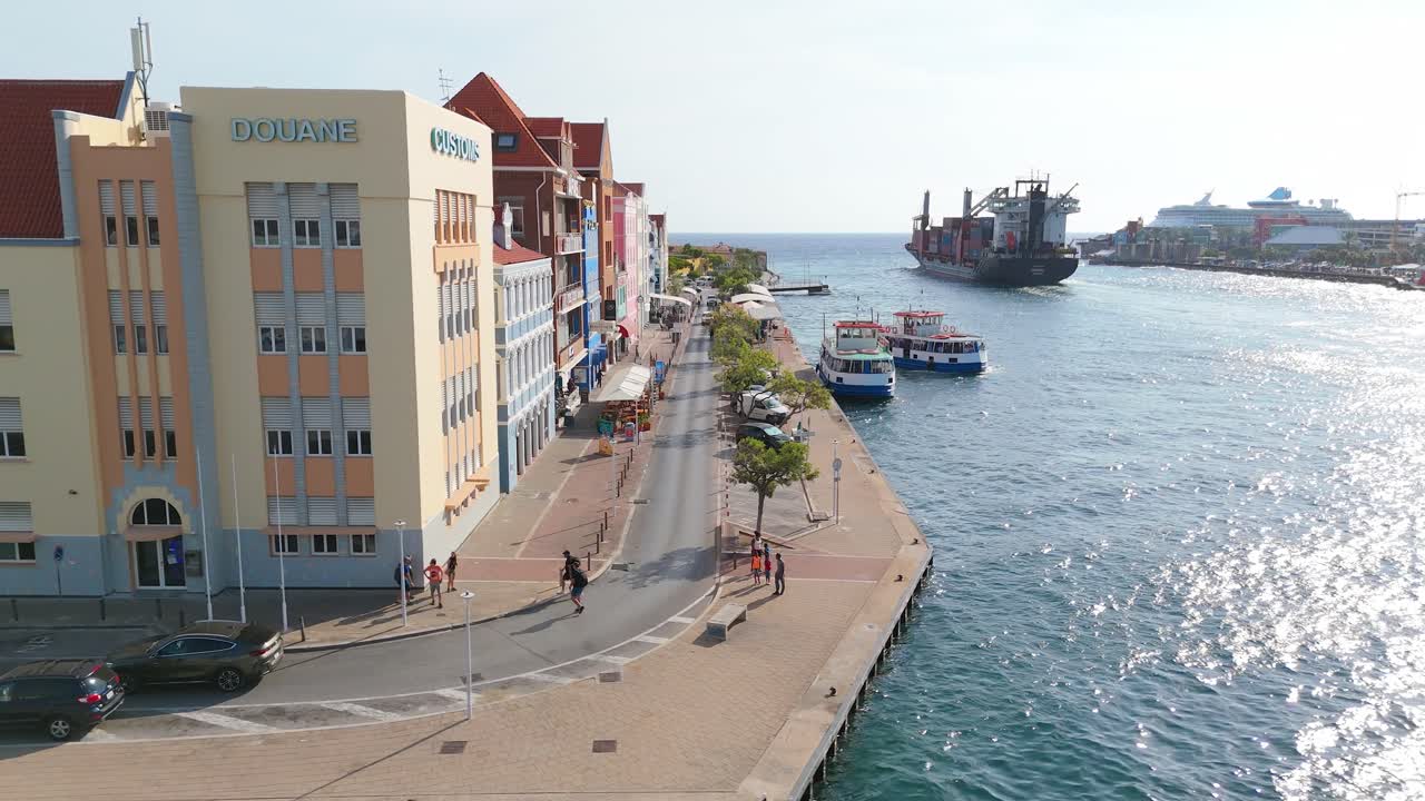 Aerial trucking pan along Punda’s colorful buildings along the coastline in Curacao, calm waters and a clear sky with ferry and cargo boat