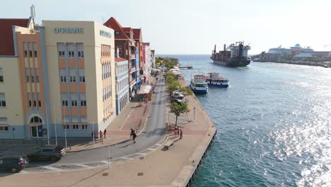 aerial trucking pan along punda’s colorful buildings along the coastline in curacao, calm waters and a clear sky with ferry and cargo boat