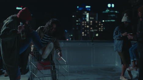 happy group of friends playing with shopping cart having fun on rooftop at night enjoying weekend celebration party