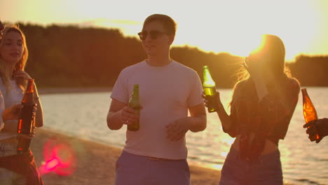 Three-girls-are-dancing-in-short-t-shirts-with-nude-waist-on-the-open-air-party-with-beer.-They-are-enjoying-the-summer-sunset-with-their-friend-on-the-beach.