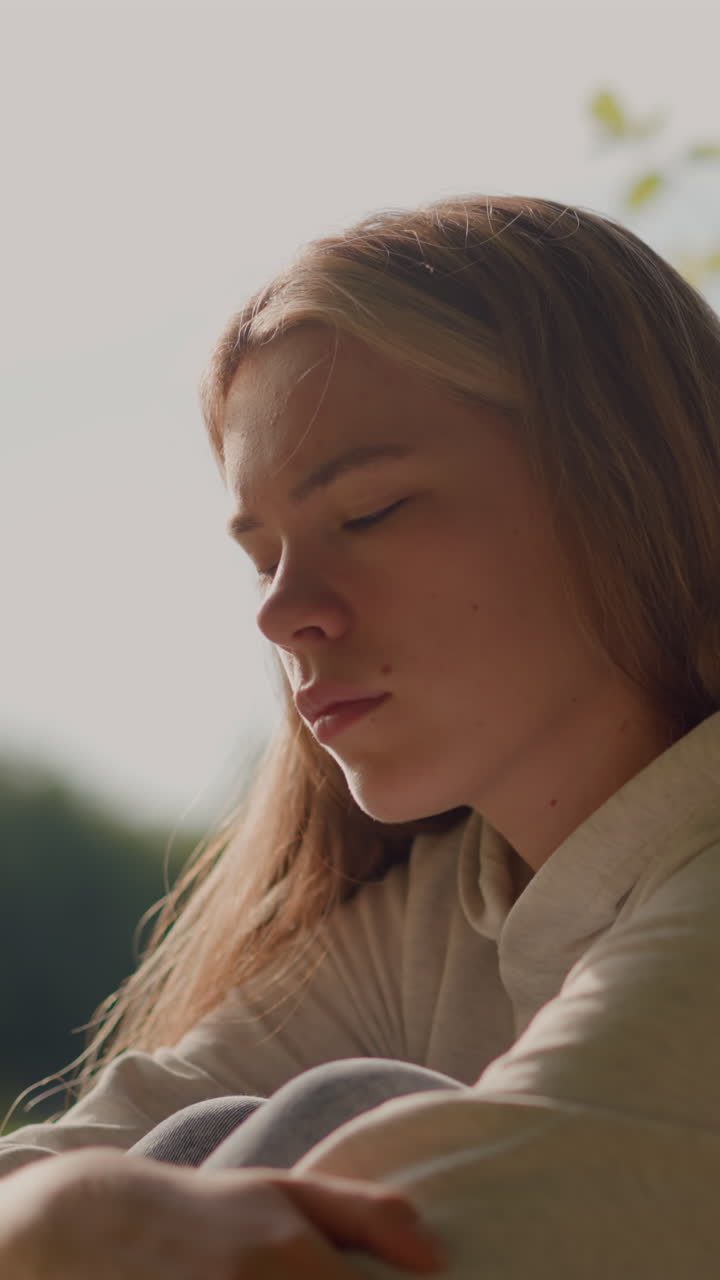 la niña se sienta en el suelo cubierto de hierba, apareciendo profundamente en pensamiento con los brazos cruzados sobre sus rodillas y la cabeza descansando suavemente en su mano, con un fondo borroso de vegetación, postes y un estadio lejano