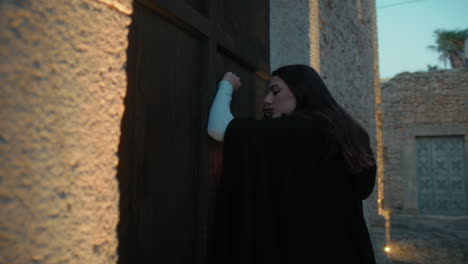 una mujer toca la puerta con una linterna por la noche en una ciudad medieval.