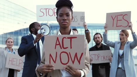 porträt einer afroamerikanerin, die act now hält"schild bei einem protest mit multiethnischen geschäftskollegen auf der straße"