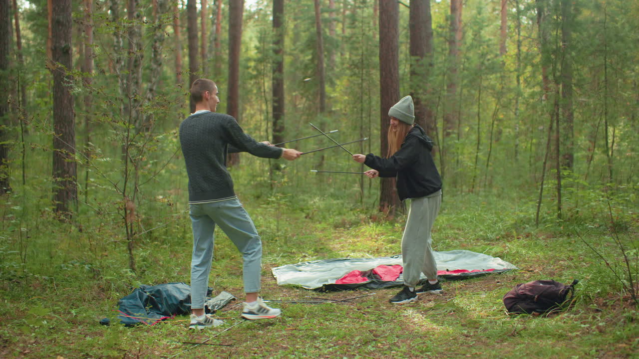 Young couple enjoy playful moment in forest while setting up tent for camping, sharing laughter and moving close for kiss, surrounded by greenery and nature in peaceful outdoor setting