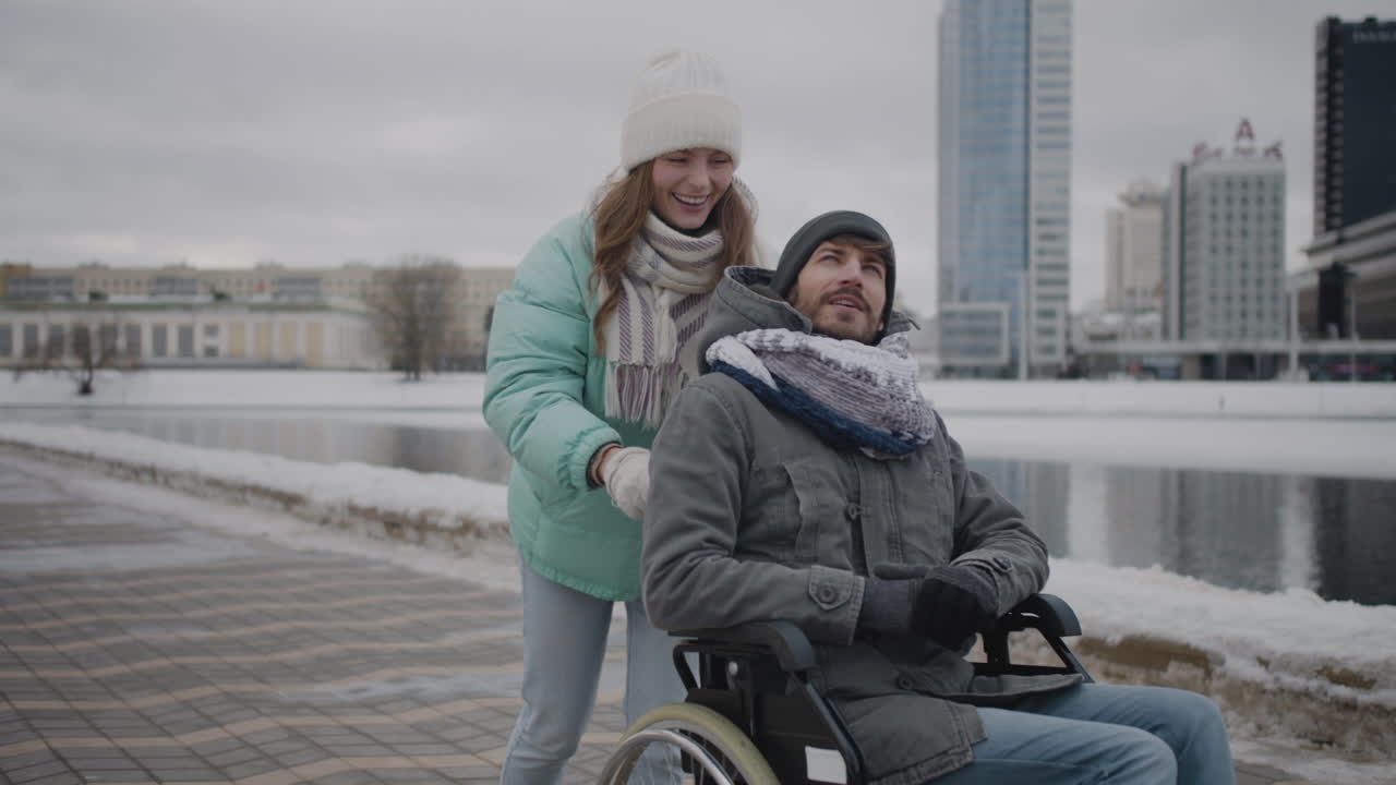 Happy Caucasian woman taking her disabled friend in wheelchair for a walk in the city. They talking together and looking at something interesting in the sky.