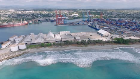 aerial wide shot of waves of caribbean sea reaching beach in front of industrial port of haina in santo domingo