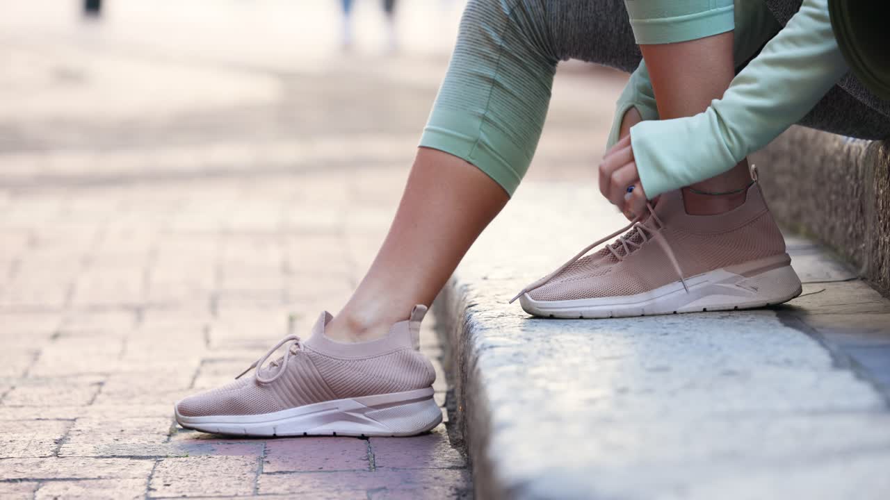 Un corridore seduto sul bordo di pietra sta allacciando i lacci delle scarpe da ginnastica rosa pallido vicino al vialetto, preparandosi a correre.