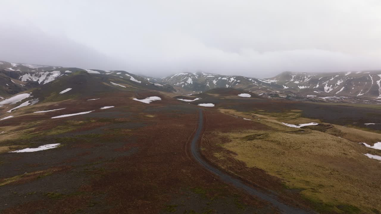 Luftbilde av et landskap med en vei som går gjennom fjell med smeltende snø, på en skyet og tåkete dag, Island