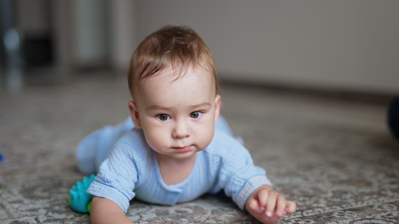 Adorable baby boy lies on the carpet with a toy in front of him. Focused little boy crawls closer to the camera. Close up.