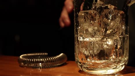 Close-up view of whiskey being poured into a glass filled with ice, with a strainer nearby.