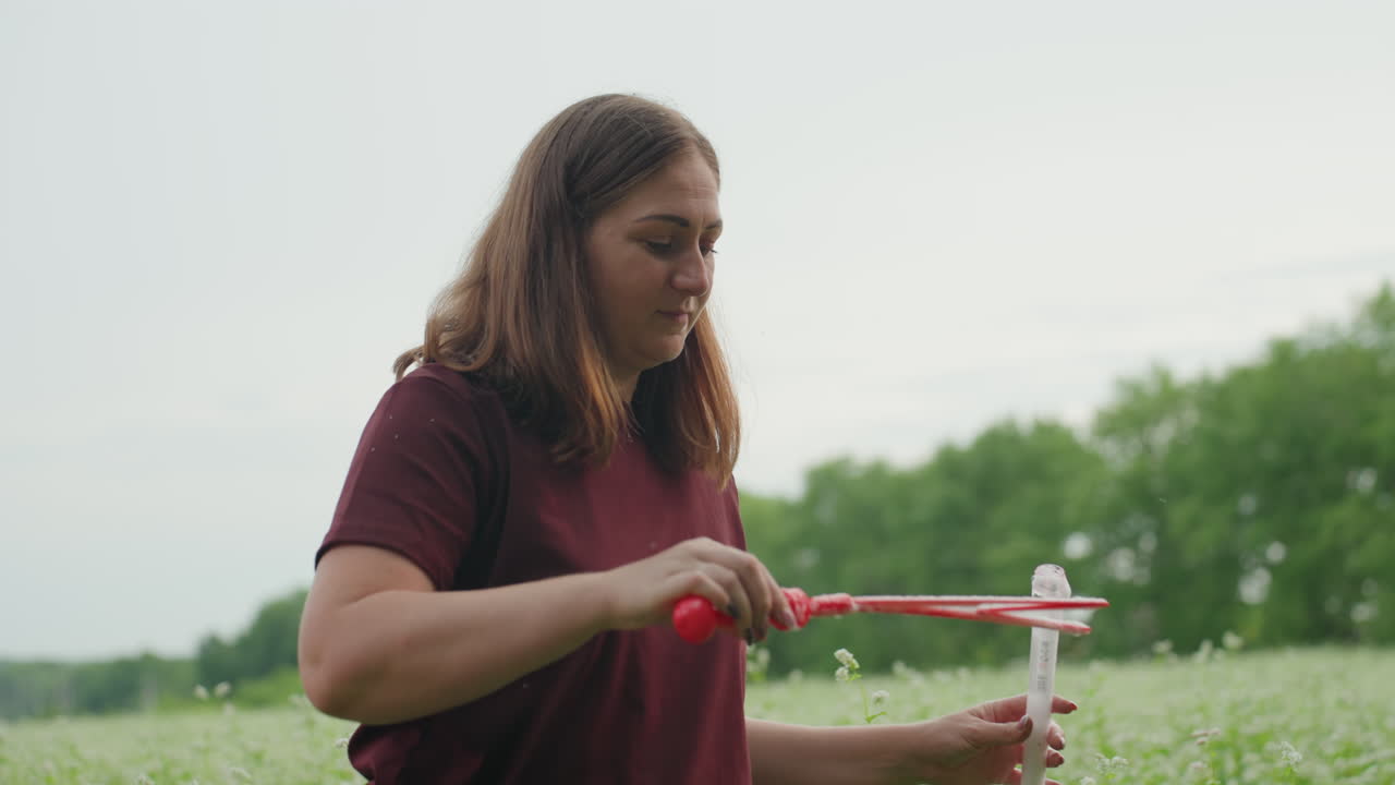Mujer caucásica soplando una varita de pompas en una pradera, camisa color burdeos creando grandes esferas de jabón iridiscentes flotando sobre un campo verde en una tarde tranquila y serena, demostración lúdica por parte de una profesora aficionada, manos