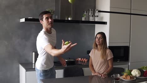 Atractiva-Pareja-Caucásica-Amorosa-Divirtiéndose-En-La-Cocina-De-Casa.-Un-Hombre-Guapo-Con-Jeans-Y-Camiseta-Blanca-Hace-Malabarismos-Con-Manzanas-Para-Impresionar-A-Su-Novia,-Ella-Lo-Abraza.-Camara-Lenta