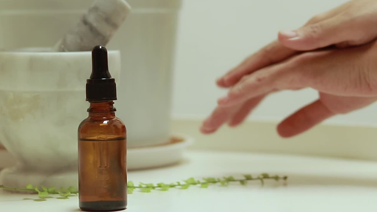 Using solution from a brown glass bottle with a dropper to moisturize hands with mortar and pestle beside plant leaves in a calm setting, concept of skincare with natural organic formulation