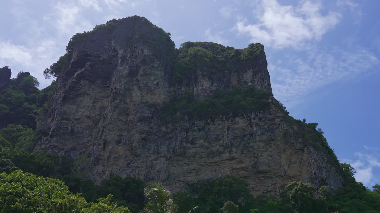 Bidikan panning lebar tebing karst batu kapur yang menjulang tinggi dikelilingi kehijauan subur di bawah langit cerah di lanskap tropis.