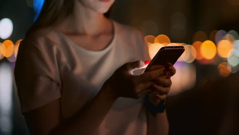 Close-up-of-a-mobile-phone-in-the-hands-of-a-girl-presses-her-fingers-on-the-screen-in-the-night-city-on-the-background-of-a-beautiful-bokeh.-Young-businessman-girl