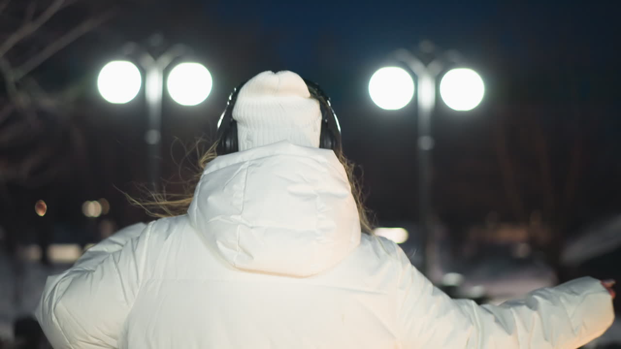 Back view of young woman in white puffer coat spins joyfully on snow covered path under bright lampposts at night with serene ambient glow and soft silhouettes of trees and park in background