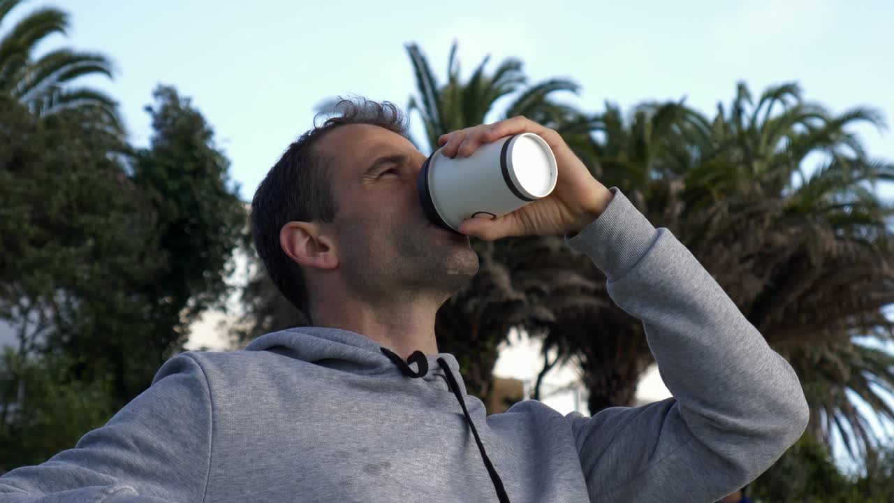 plano medio de un joven caucásico tomando café sentado en un banco en la playa en auckland, nueva zelanda