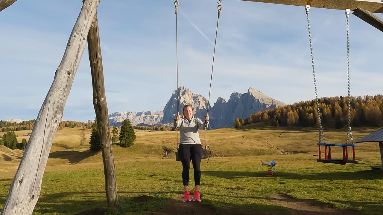mujer aventurera se balancea en un conjunto de madera con grandes campos de hierba exuberante y picos escarpados de las dolomitas detrás