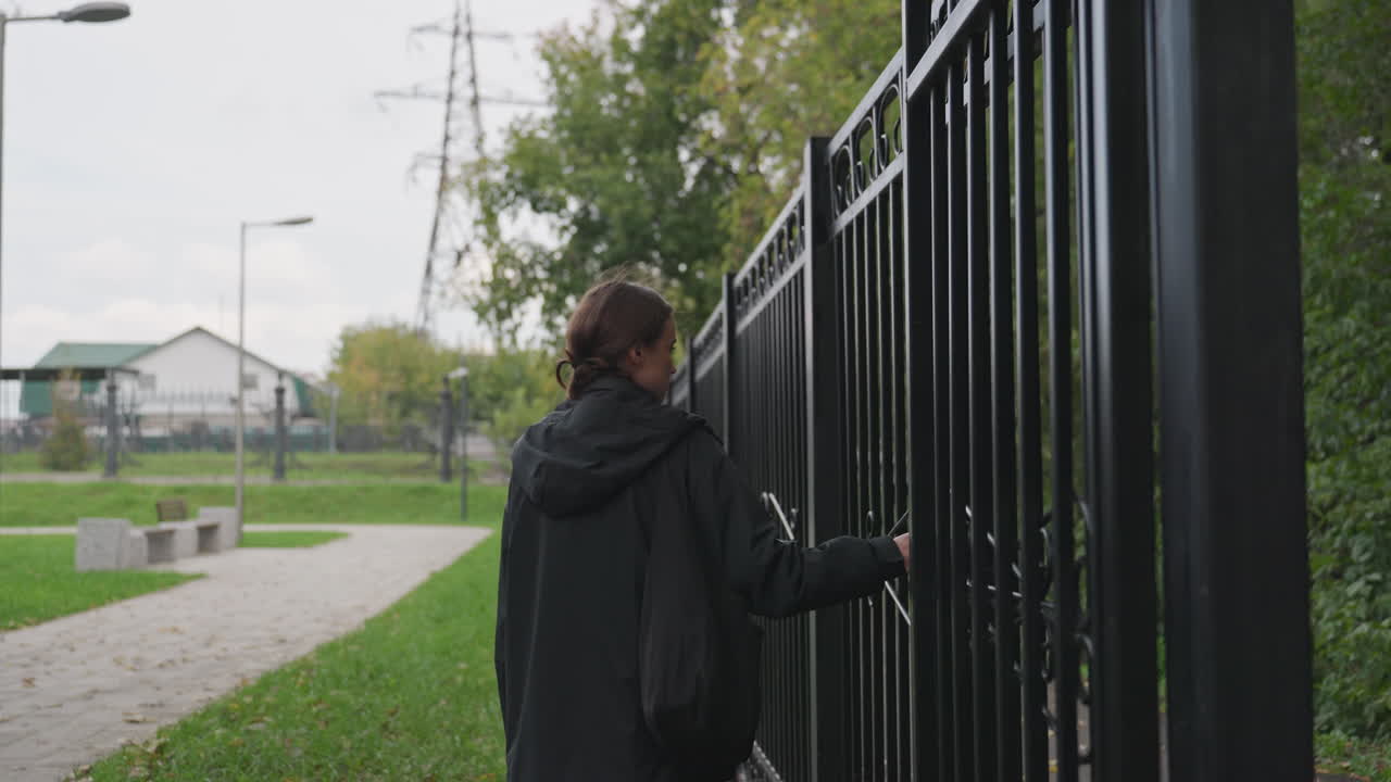 Donna che passeggia in un parco di periferia, donna che tocca delicatamente un cancello mentre cammina sotto un cielo nuvoloso, donna con una giacca con cappuccio che cammina tranquillamente e si appoggia alle ringhiere in un tranquillo parco
