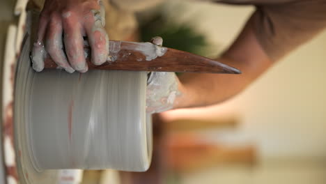 vertical potter hands using wooden tool shaping clay vase on workshop turntable