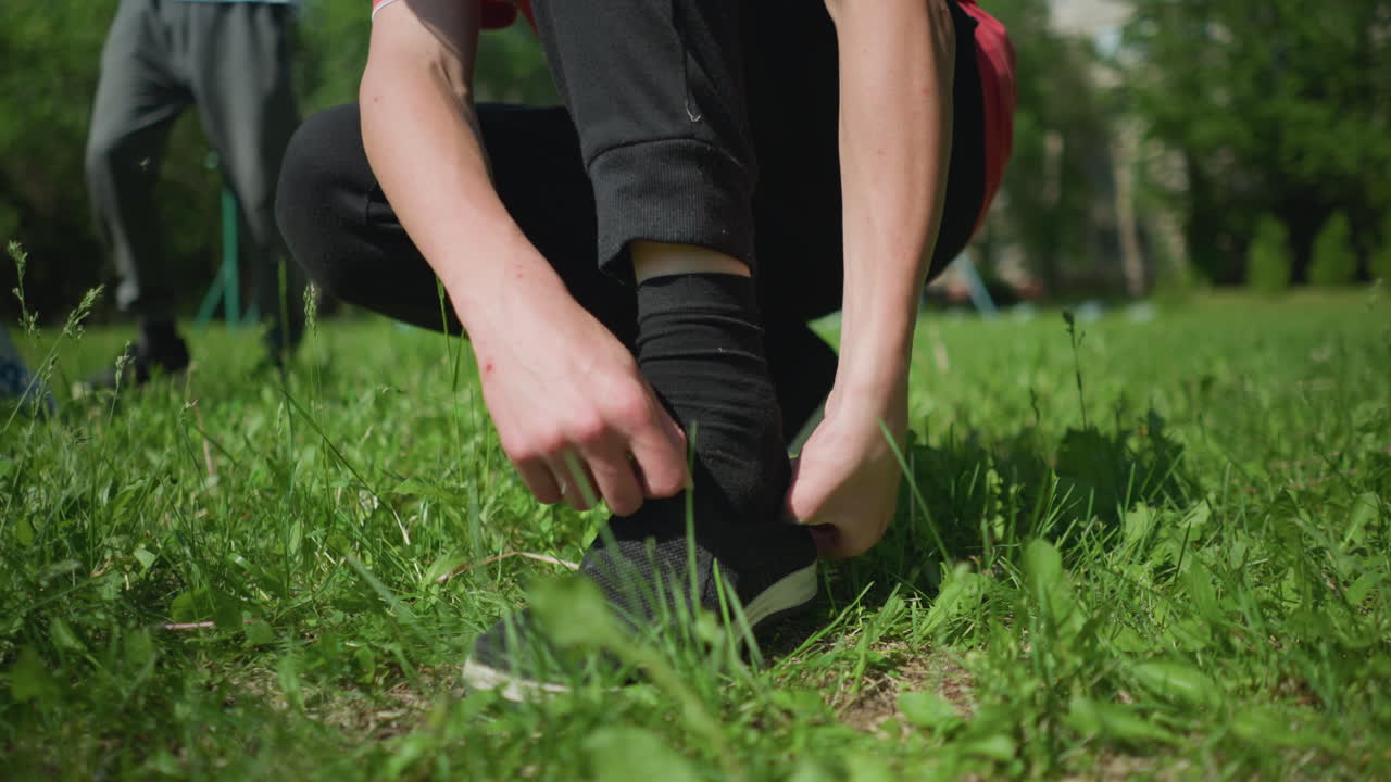 Close-up of someone adjusting and wearing their shoes while kneeling on a grassy field, with another person visible moving in the background