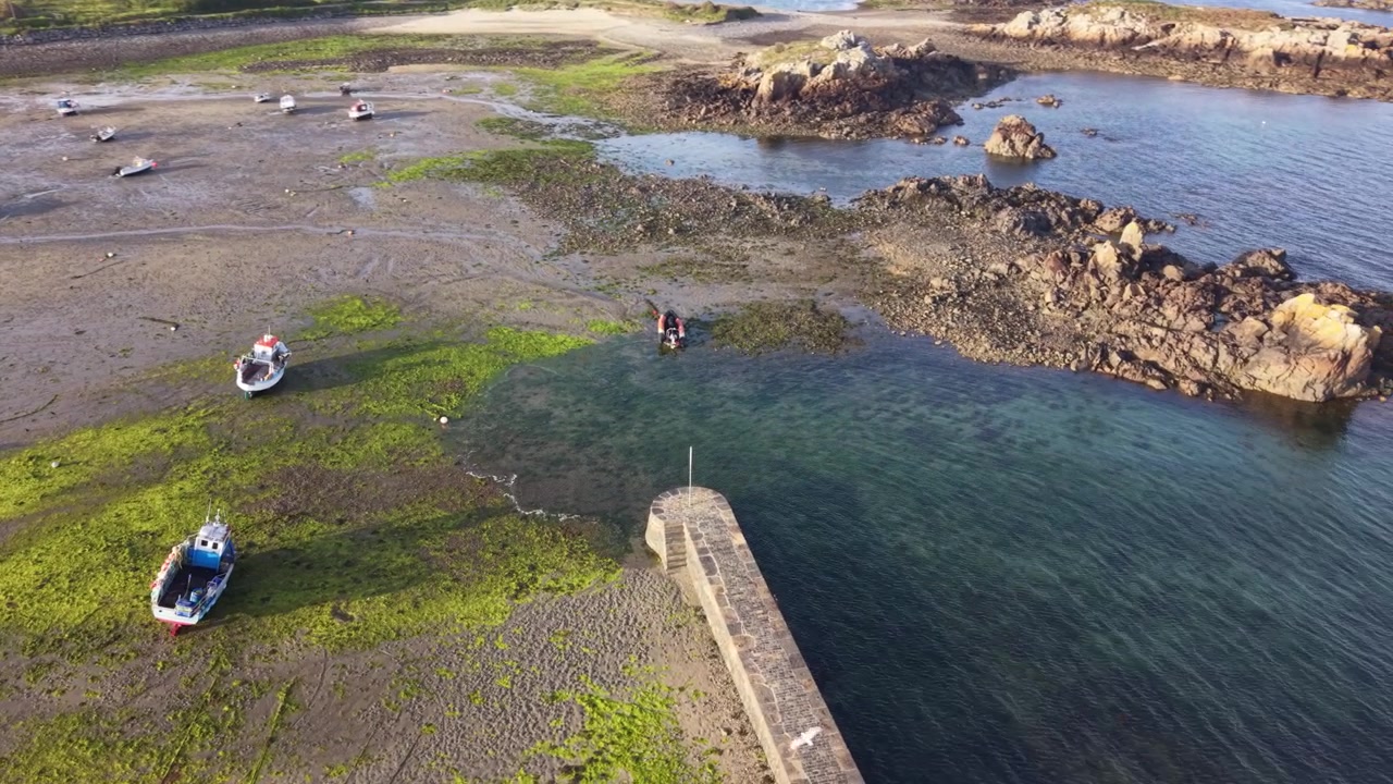 Bordeaux'n satama, Guernsey, Kanaalisaaret. Dramaattista ympyränmuotoista dronekuvaa merimuurin yllä, kristallinkirkkaan veden, ohi lentävien lintujen ja hiekkaisella merenpohjalla kuivuvien pienten veneiden kera myöhäisen iltapäivän auringossa.