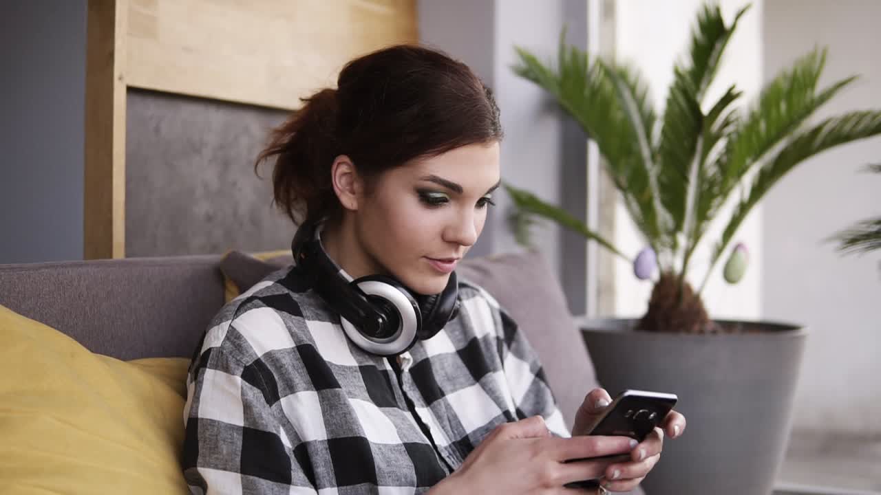 una hermosa mujer morena se sienta en un sofá con almohada amarilla. sosteniendo el teléfono móvil en las manos, desplazándose y sonriendo. en el interior