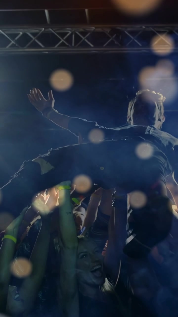 Video verticale: Uomo con le braccia alzate, sollevato dalla folla durante un concerto sotto le calde luci del palco, per divertimento.