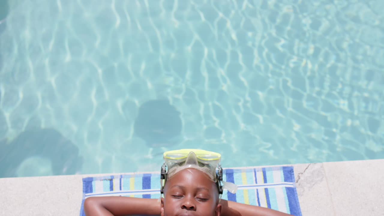 High angle of happy african american boy in goggles sunbathing by pool, copy space, slow motion