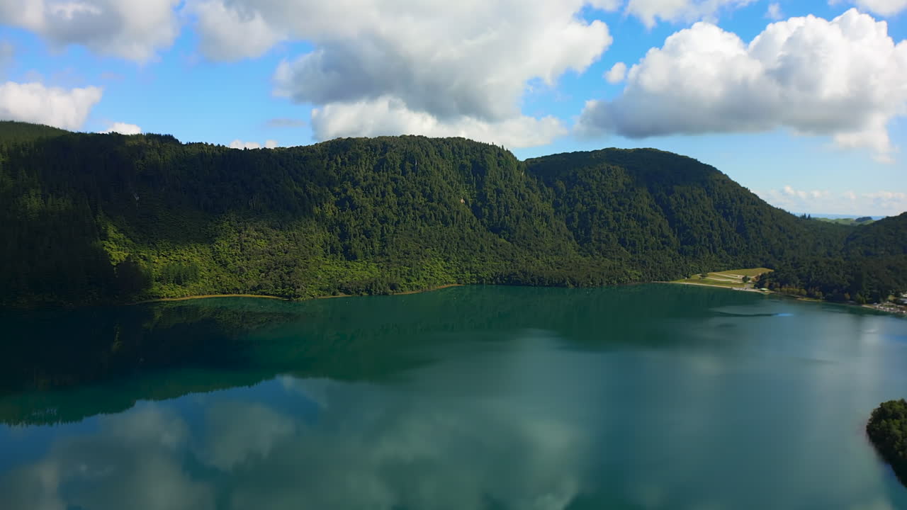 Lush green hills reflect on Blue Lake in Rotorua, under a bright blue sky with fluffy clouds
