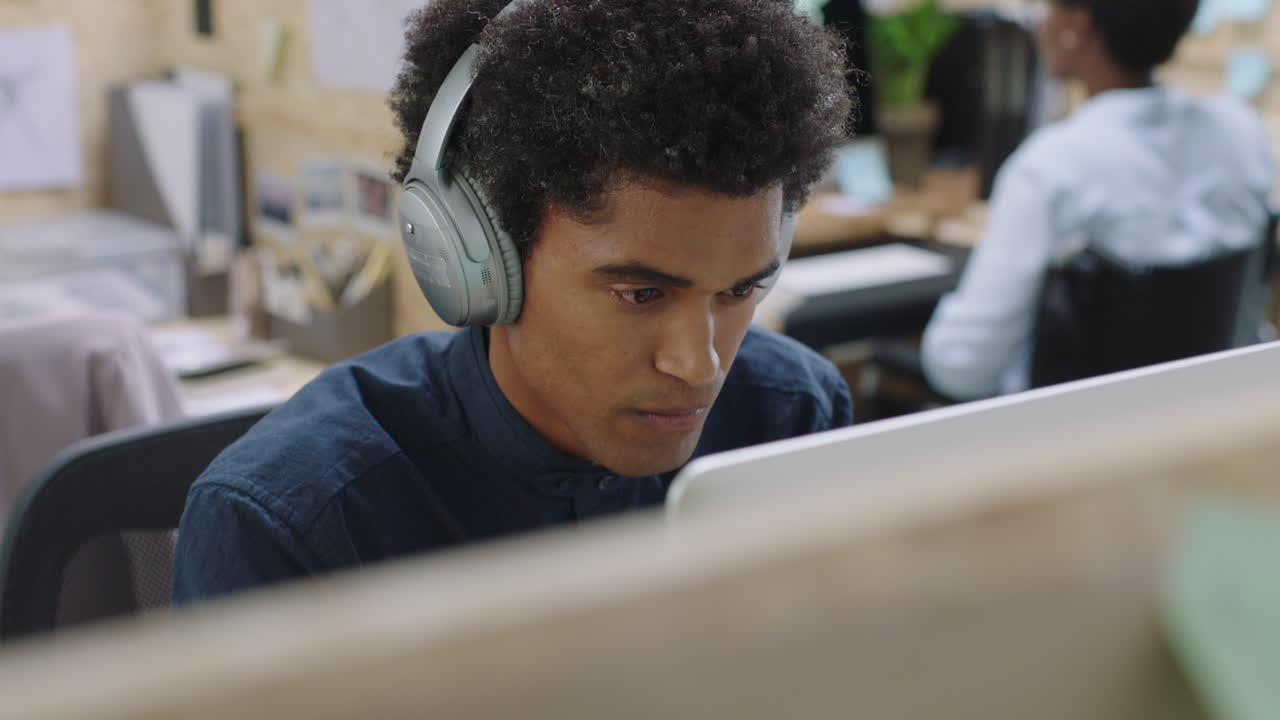 young mixed race businessman using computer working on business project browsing online listening to music brainstorming ideas wearing headphones in relaxed office workplace