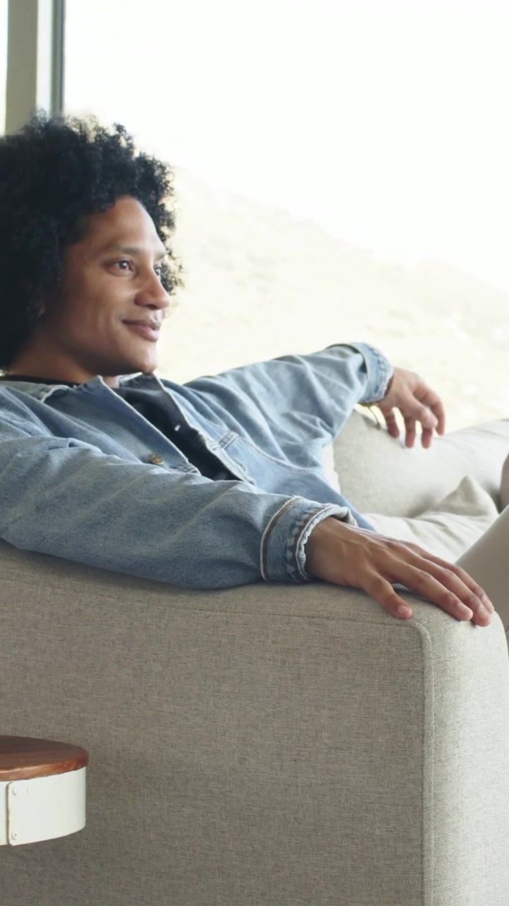 Video verticale: Uomo con camicia di jeans che si raddrizza e sorride, reagendo a una finestra luminosa mentre è sul divano.