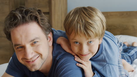 close-up view of happy father and his cute little son hugging him from back, while they lying on the bed and looking at camera in the morning