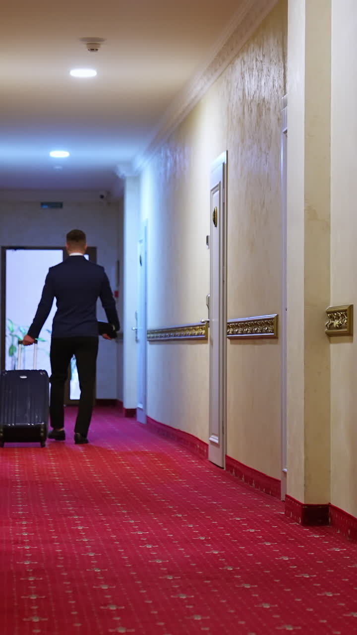 Back view of businessman walking with suitcase in hotel corridor. Modern interior with white walls, doors and red carpet. Vertical video