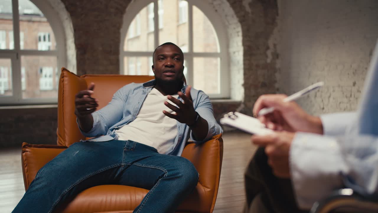 A man with Black skin in a blue shirt and jeans sits on a light brown one with a professional psychologist who holds a tablet and a pen in his hands