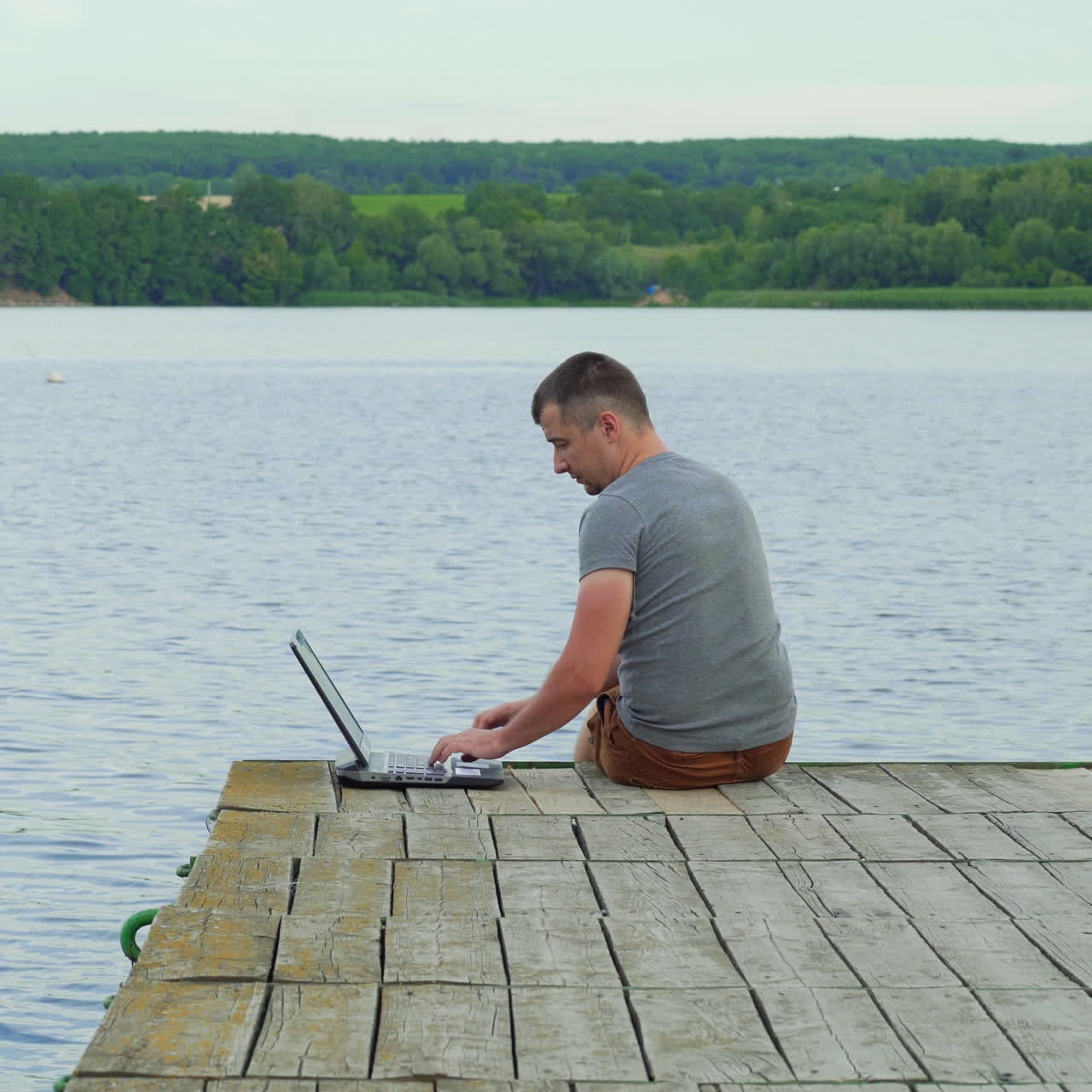 Handsome man sitting at the edge of the bridge finishes typing on a laptop and enjoys the natural background. Work with pleasure outdoors near the river